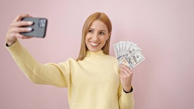 Sarışın genç bir kadın, izole edilmiş pembe arka planda elinde dolarlarla akıllı telefonuyla selfie çekiyor.