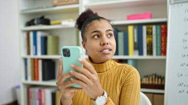 Genç Afrikalı Amerikalı bayan öğrenci kütüphanede akıllı telefon kullanıyor.