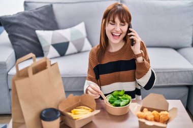 Akıllı telefondan konuşan genç bir kadın yemek yerken evde yemek yiyor.