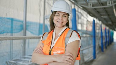 Orta yaşlı İspanyol kadın müteahhit inşaat alanında kollarını kavuşturup gülümsüyor.