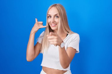 Young caucasian woman standing over blue background smiling doing talking on the telephone gesture and pointing to you. call me. 