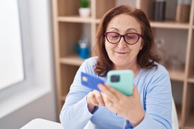 Senior woman using smartphone and credit card sitting on table at home