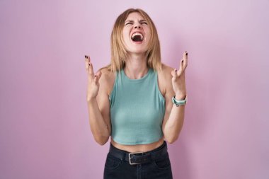 Blonde caucasian woman standing over pink background crazy and mad shouting and yelling with aggressive expression and arms raised. frustration concept. 