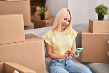 Genç sarışın kadın akıllı telefon kullanıyor. Yeni evinde yerde oturuyor.