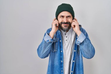 Dövmeli, yün şapkalı, parmaklarıyla kulaklarını kapatan, gürültülü müzik sesi için rahatsız edici ifadeleri olan genç İspanyol bir adam. sağır kavramı. 