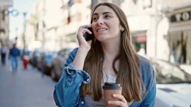 Young beautiful hispanic woman talking on smartphone drinking coffee at street