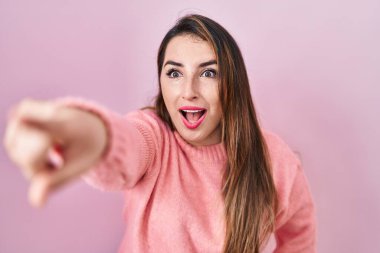 Young hispanic woman standing over pink background pointing with finger surprised ahead, open mouth amazed expression, something on the front 