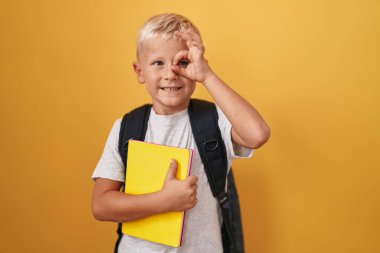 Küçük beyaz çocuk, öğrenci çantası takıyor ve elinde kitap tutuyor. Gülümsüyor. Tamam. Elleri gözlerinde, parmaklarından bakıyor. 