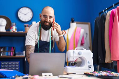 Young bald man tailor talking on smartphone using laptop at clothing factory