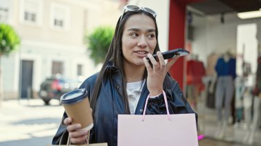 Güzel İspanyol bir kadın elinde alışveriş poşetleri ve sokakta kahve tutarak akıllı telefonuyla sesli mesaj yolluyor.