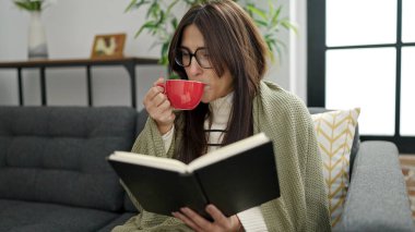Young beautiful hispanic woman reading book and drinking coffee sitting on sofa at home