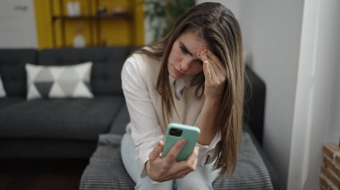 Genç, güzel, İspanyol bir kadın evde endişeli bir ifadeyle akıllı telefon kullanıyor.