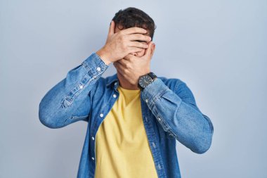 Young hispanic man standing over blue background covering eyes and mouth with hands, surprised and shocked. hiding emotion 