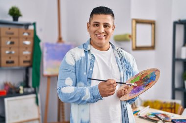 Sanat stüdyosunda palet ve boya fırçasıyla gülümseyen genç Latin sanatçı.