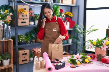 Young chinese woman florist talking on smartphone holding gift bag at flower shop