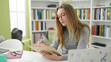 Kütüphane üniversitesinde masaya oturan beyaz kadın öğrenci belgeye yazı yazıyor.