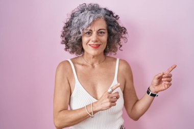 Pembe arka planda gri saçlı orta yaşlı bir kadın gülümsüyor ve kameraya bakıyor.. 