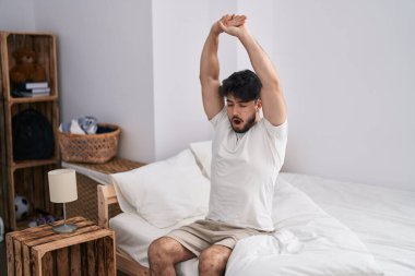 Young hispanic man waking up stretching arms at bedroom