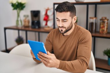 Young hispanic man using touchpad sitting on table at home
