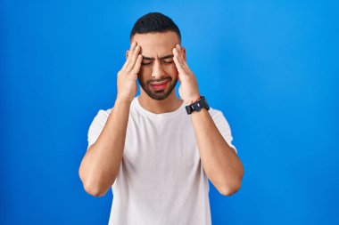 Young hispanic man standing over blue background with hand on head, headache because stress. suffering migraine. 