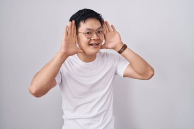 Young asian man standing over white background smiling cheerful playing peek a boo with hands showing face. surprised and exited 