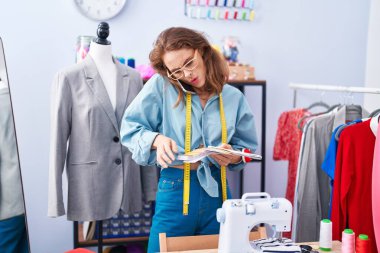 Young beautiful hispanic woman tailor talking on smartphone looking color test at tailor shop