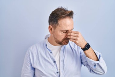 Orta yaşlı, beyaz bir adam mavi arka planda dikiliyor. Burnu ve gözleri ovuşturmaktan yorulmuş, yorgunluk ve baş ağrısı çekiyor. Stres ve hüsran kavramı. 