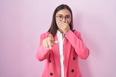 İş kıyafetleri ve gözlük takan genç İspanyol kadın sana gülüyor, eli ağzında kamerayı işaret ediyor, utanç ifadesi var. 