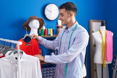 Young hispanic man tailor holding t shirt on rack at sewing studio