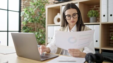 Young beautiful hispanic woman business worker using laptop reading document at office