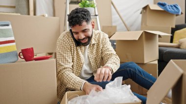Genç İspanyol adam akıllı telefondan konuşuyor. Yeni evdeki karton kutuları açıyor.