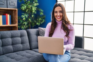 Genç İspanyol kadın evdeki koltukta dizüstü bilgisayar kullanıyor.