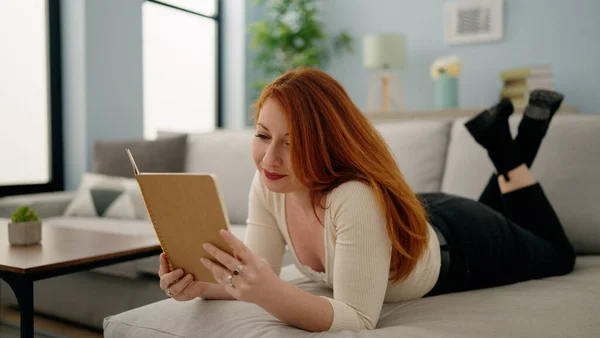 Young Redhead Woman Reading Book Lying Sofa Home — Foto Stock