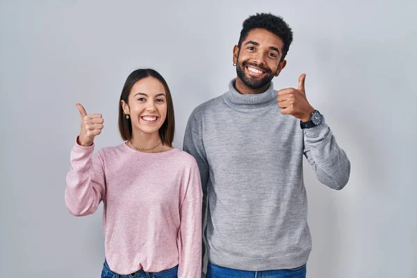 stock image Young hispanic couple standing together doing happy thumbs up gesture with hand. approving expression looking at the camera showing success. 