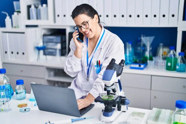 Young Hispanic Woman Scientist Talking Smartphone Using Laptop Laboratory — Stock fotografie
