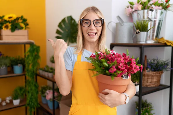 年轻的高加索女人在花店工作时 拿着植物尖着大拇指朝天微笑着张开嘴 — 图库照片