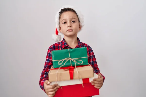 クリスマスの帽子をかぶっている小さなヒスパニック系の少年は 顔に深刻な表情でリラックスしたプレゼントを保持する カメラを見ているだけで — ストック写真