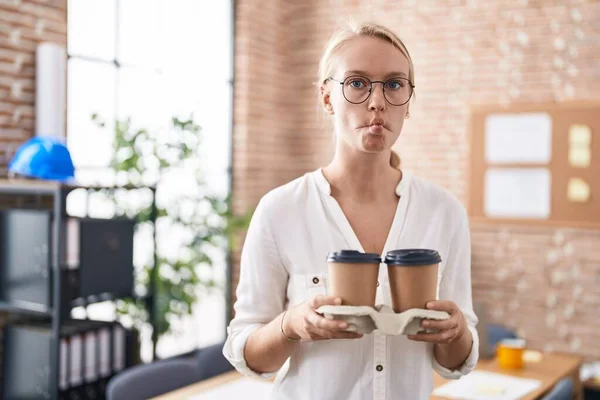 stock image Young caucasian woman working at the office holding coffee cups making fish face with mouth and squinting eyes, crazy and comical. 