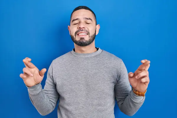 Hombre Hispano Pie Sobre Fondo Azul Gesto Dedo Cruzado Sonriendo —  Fotos de Stock