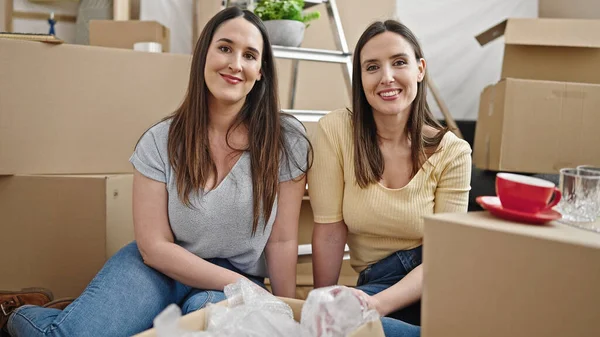 İki kadın yeni evinde yerde kendinden emin bir şekilde gülümsüyor.