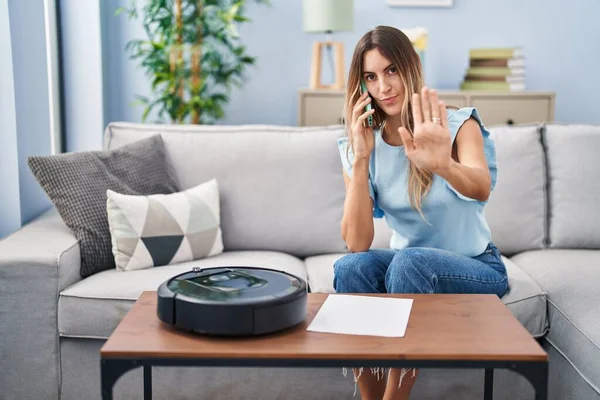 Young hispanic woman sitting at home by vacuum robot speaking on the phone with open hand doing stop sign with serious and confident expression, defense gesture