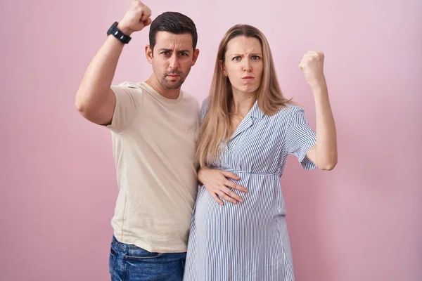 ピンク色の背景に立っている赤ちゃんを期待している若いカップルは怒りながら怒りと怒りを叫びながら拳を上げる怒っている 怒りと攻撃的な考え方 — ストック写真