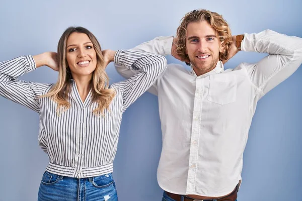 Mavi arka planda duran genç çift rahatlıyor ve esniyor, kolları ve elleri başlarının arkasında mutlu bir şekilde gülümsüyor. 