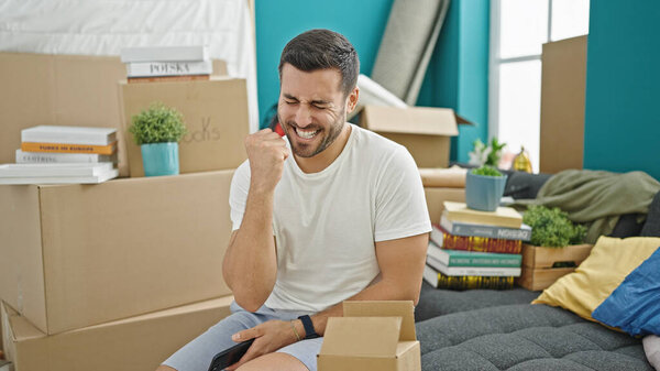 Молодой латиноамериканец держит смартфон с выражением победителя в новом доме