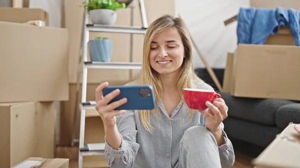 若いですブロンド女性使用スマートフォン飲料コーヒーで新しい家 — ストック写真