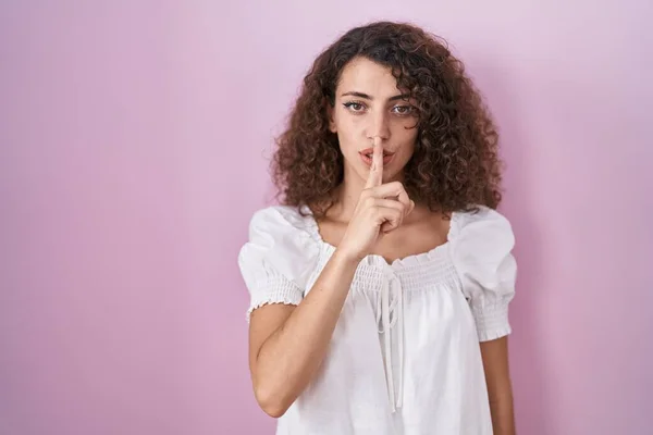 头发卷曲的西班牙裔女人站在粉红的背景上 要求安静 手指放在嘴唇上 沉默和秘密概念 — 图库照片