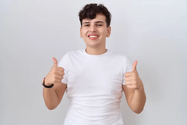 カジュアルな白いTシャツの成功の印を身に着けている若い非バイナリの人は手で肯定的なジェスチャーをする 親指を笑顔と幸せ 陽気な表情と勝者のジェスチャー — ストック写真