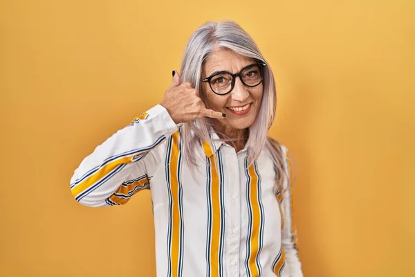 中年妇女 白发苍苍地站在黄色的背景上 戴着眼镜 微笑着 用手和手指做着电话手势 就像在电话里说话一样 交流概念 — 图库照片