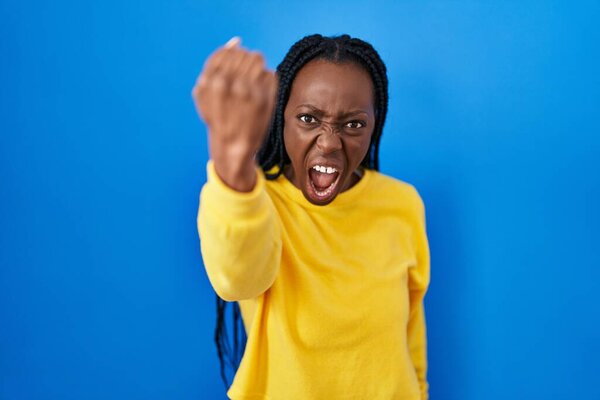 Beautiful black woman standing over blue background angry and mad raising fist frustrated and furious while shouting with anger. rage and aggressive concept. 