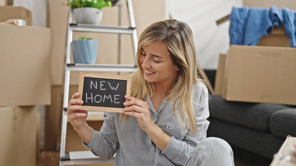 Young blonde woman holding blackboard sitting on floor at new home